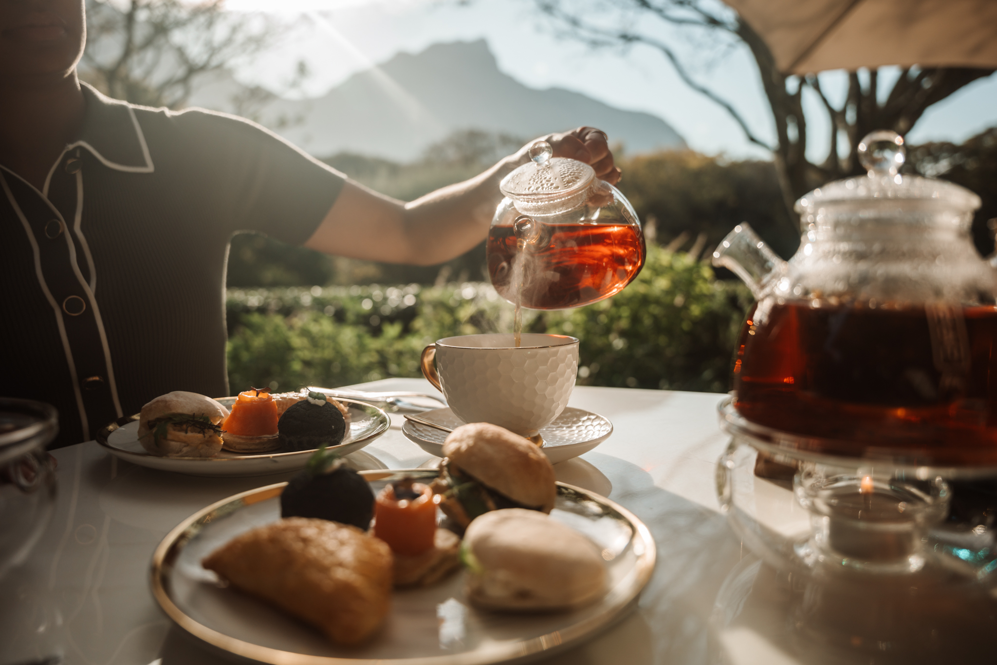 Person pours red tea from a glass teapot into a white textured cup on an outdoor table with pastries and a second teapot nearby, sunlit garden setting.