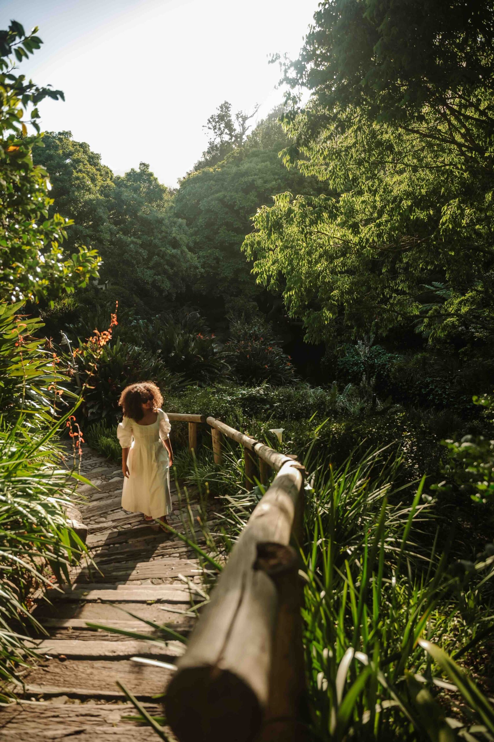 Woman in a light dress walking down a wooden steps path surrounded by lush green plants in a garden sunlit scene