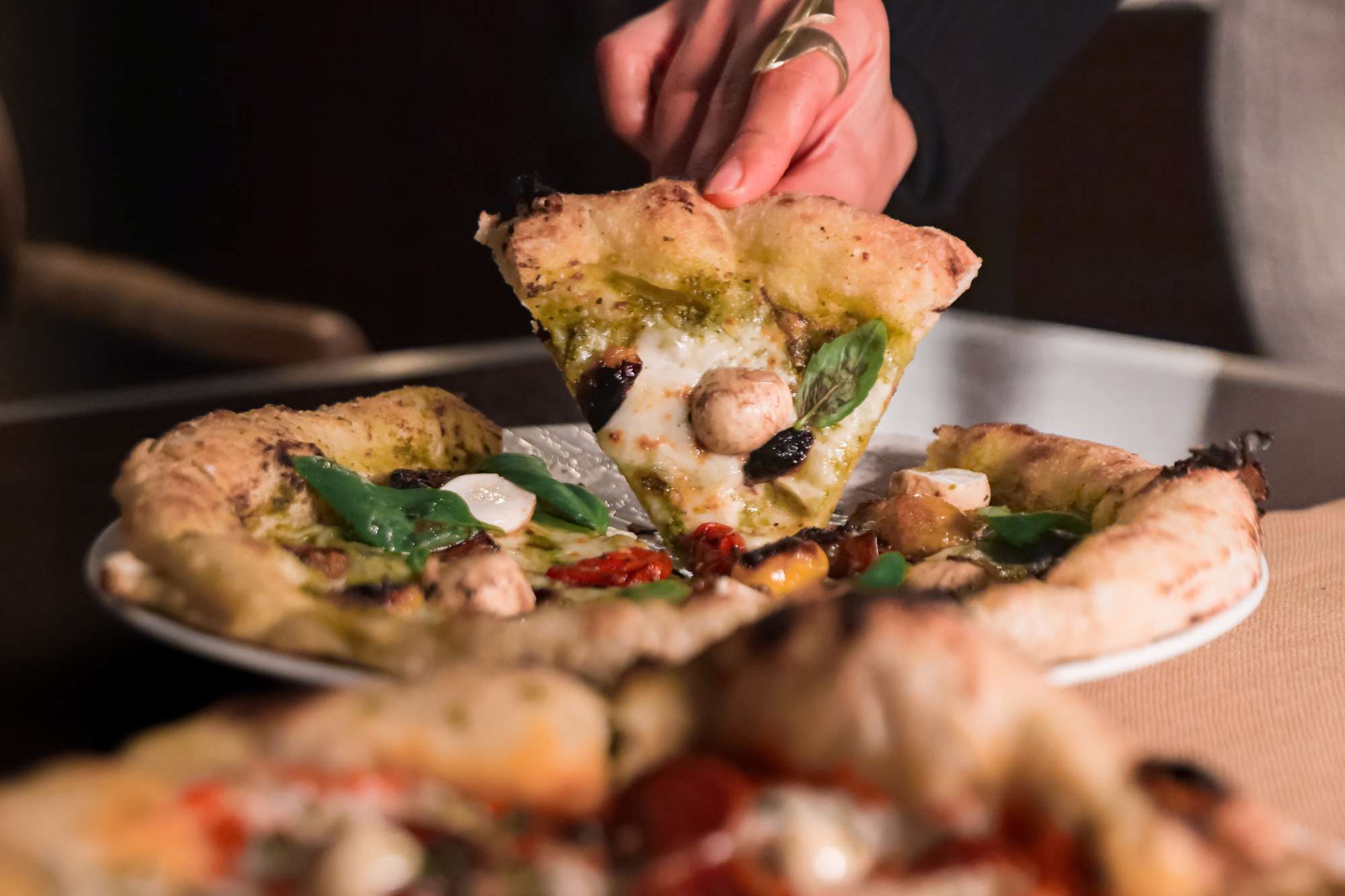 1 | Vineyard Hotel Person holding a slice of chunky veggie pizza with basil leaves and melted cheese over a plate.