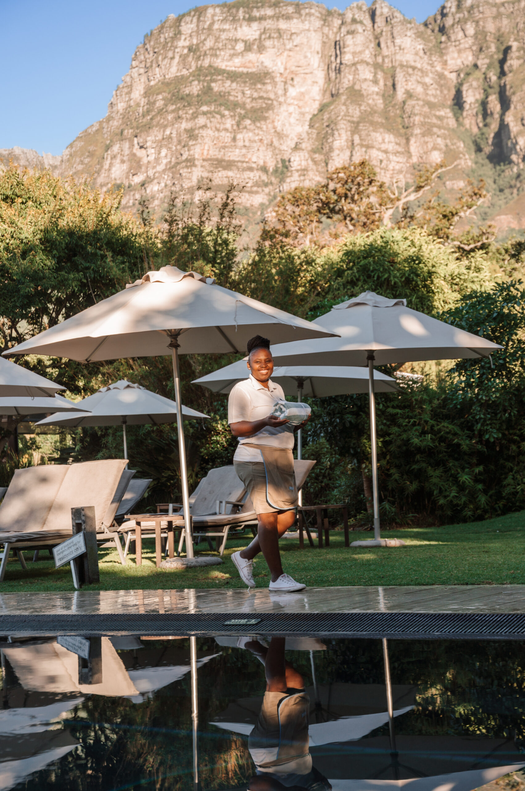 Smiling woman in light clothing carrying towels near a pool under beige umbrellas with a rocky mountain backdrop behind her.