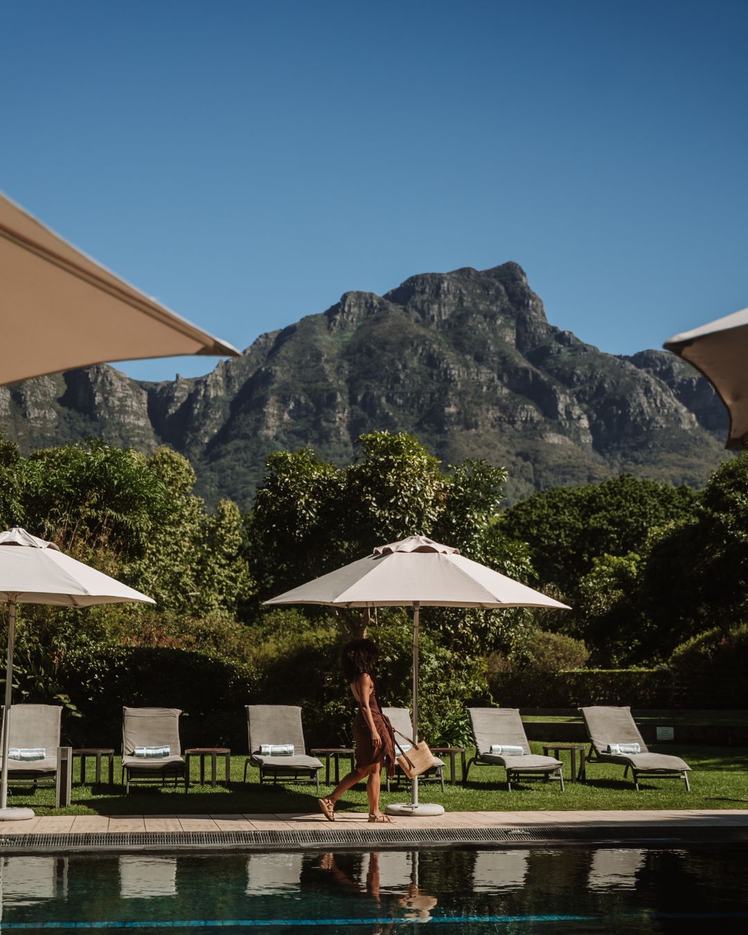 Media New Section | Vineyard Hotel Poolside scene with white umbrellas and lounge chairs, a person in a brown dress walking along the pool, with mountains in the distance under a blue sky.