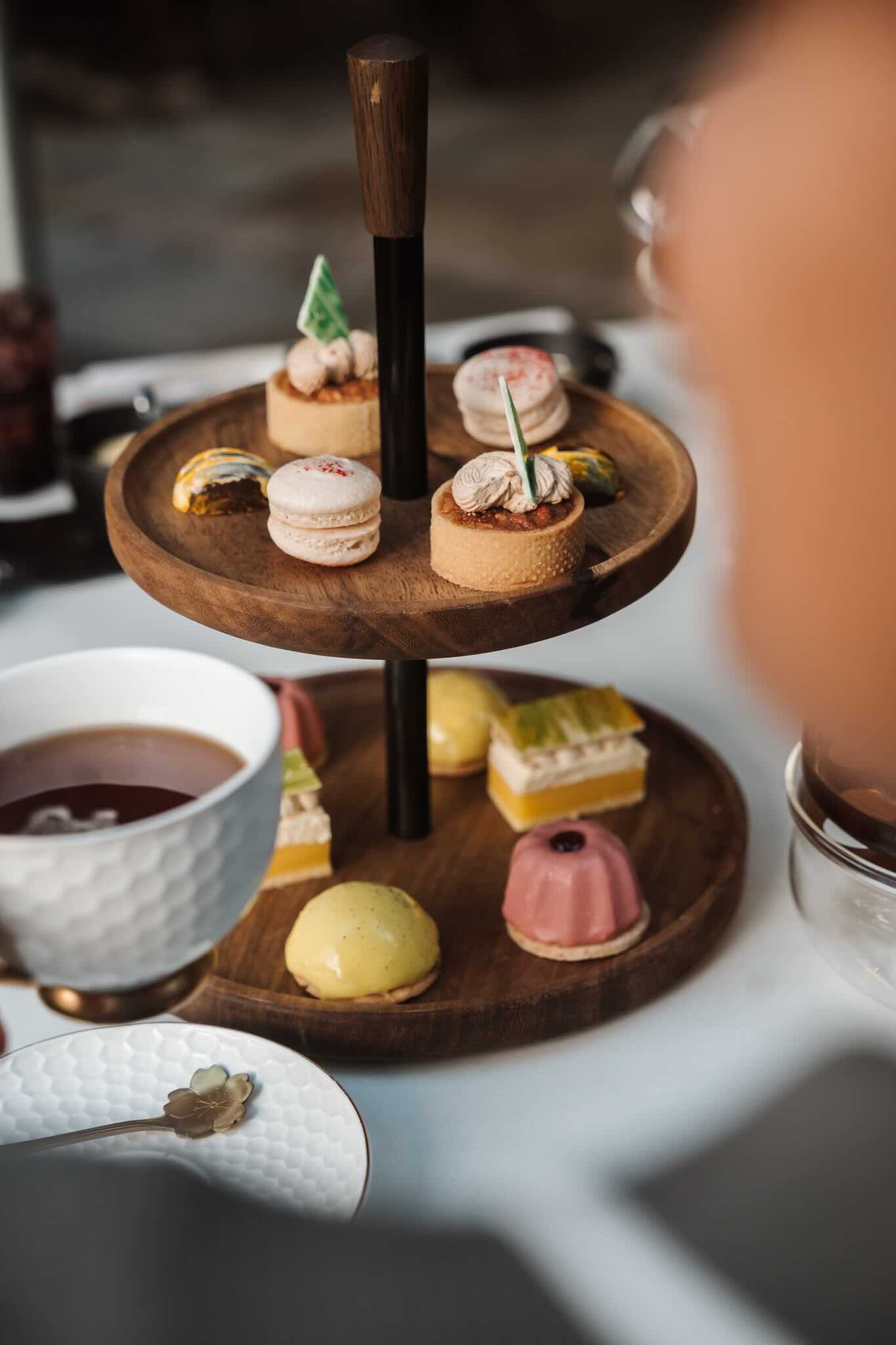 VINEYARD-002 | Vineyard Hotel Assorted desserts on a two-tier wooden stand, including macarons and small cakes, with a cup and spoon in the foreground.
