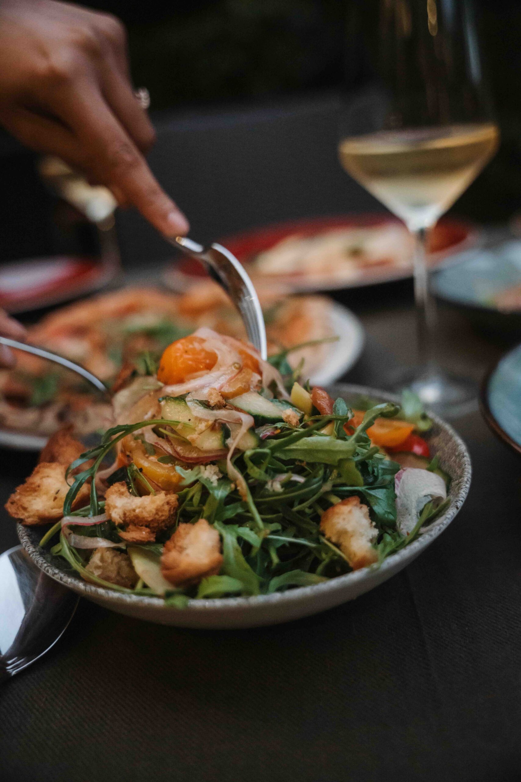 Close-up of a plated salad with greens, croutons, and sliced vegetables, as a hand slices with a knife in the background; a wine glass nearby.