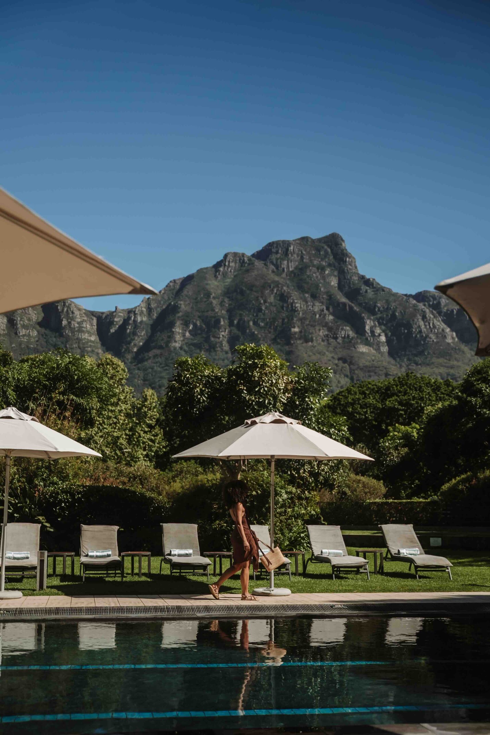 VINEYARDHOTEL-60 | Vineyard Hotel Poolside area with white umbrellas, lounge chairs, and a person walking along the pool edge, mountains in the distance under a blue sky.