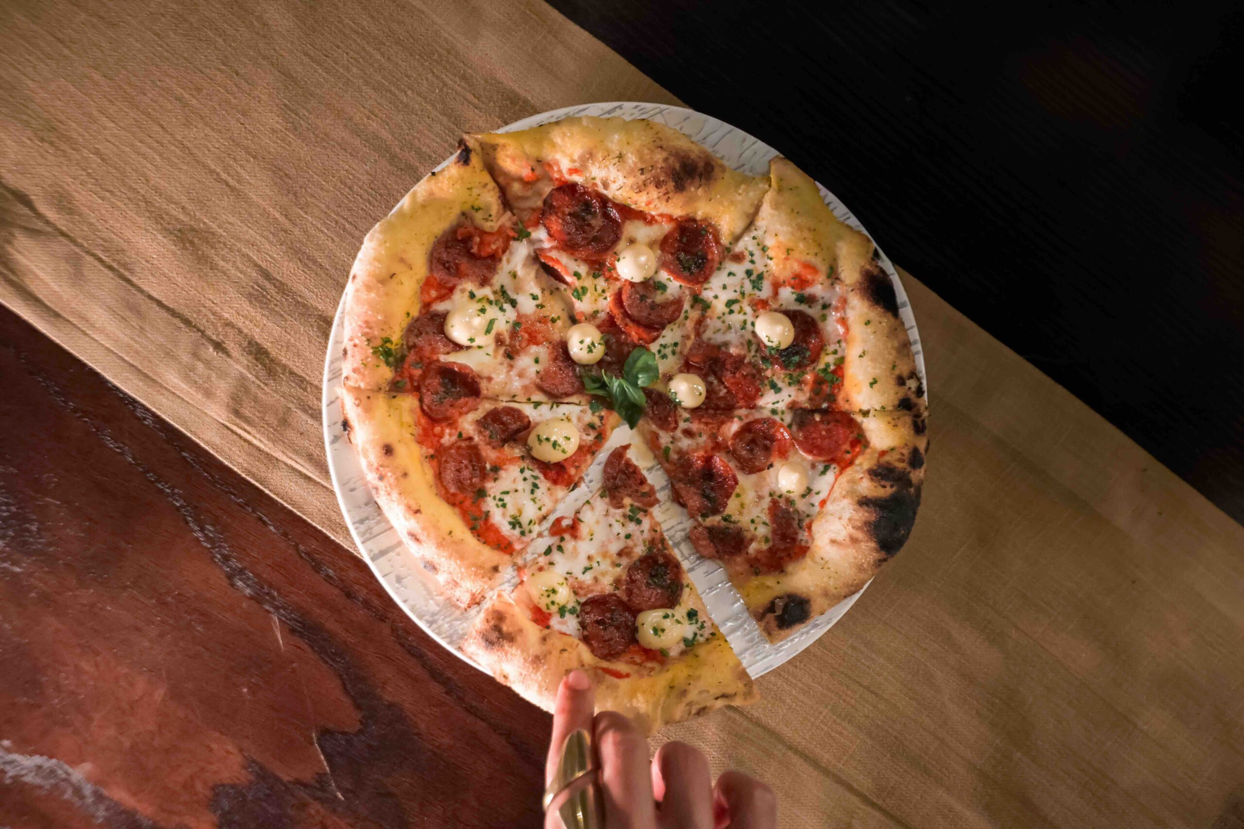 Overhead view of a pepperoni pizza on a white plate, with melted cheese, herbs, and a hand holding a fork and knife at the bottom edge of the frame over a wooden table.