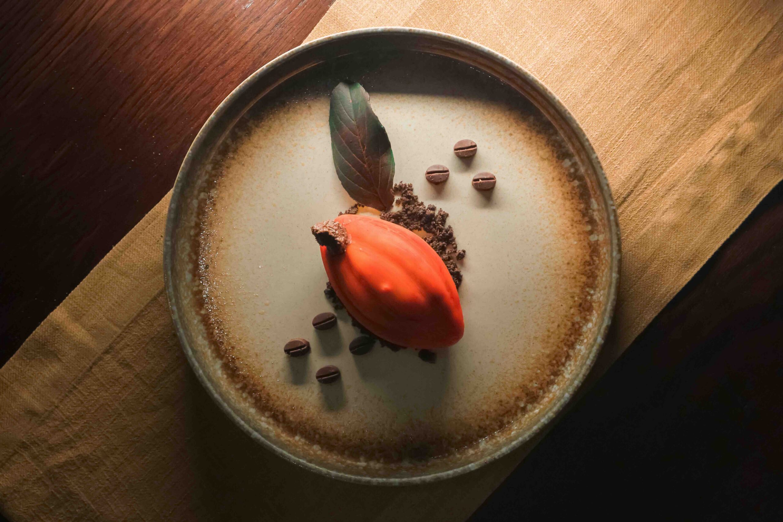 Dessert plate with orange scoop of sorbet, chocolate crumbs, a basil-like leaf, and scattered chocolate chips on a speckled ceramic dish.