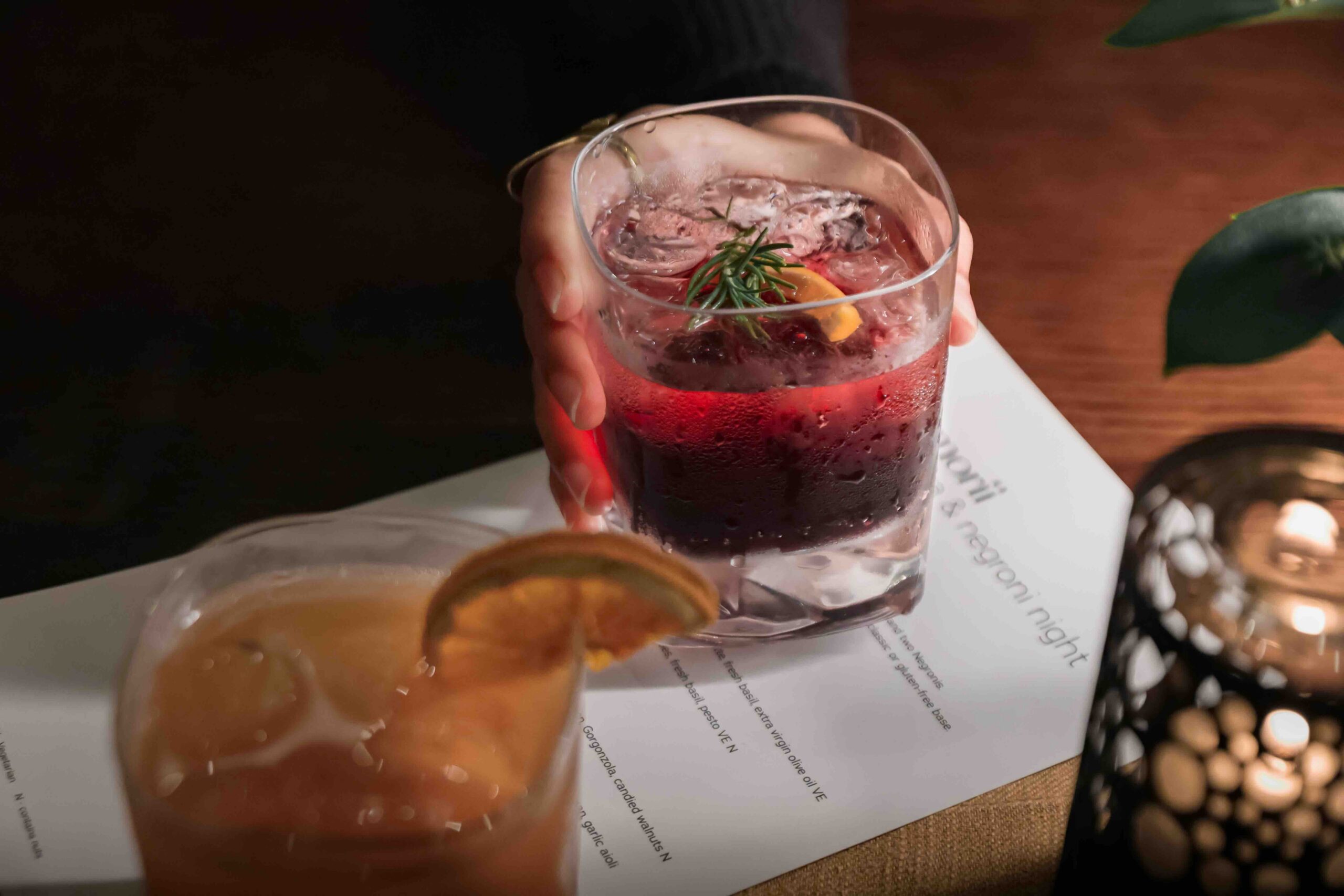 Hand holding a glass of red berry cocktail with ice, rosemary sprig and orange slice.