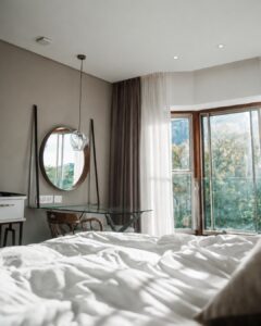 Bright modern bedroom corner with a glass desk, round mirror, and hanging glass pendant light, near large windows with sheer curtains and a view of greenery outside.