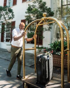 Porter in a white shirt pushes a gold-toned luggage cart with a black suitcase outside a white building with plants nearby.
