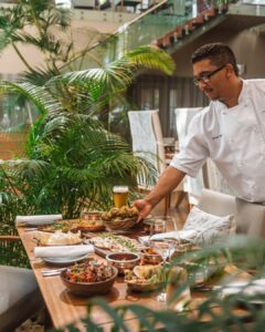 Chef in a white coat serves a long wooden table piled with appetizers and a glass of beer in a tropical indoor garden setting.