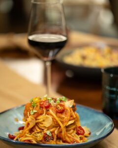 Plate of spaghetti with tomato sauce, cherry tomatoes, and herbs topped with crispy parmesan chips, against a blurred wine glass backdrop