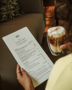 Person holding a café menu titled 'Long Café' with a frothy glass of beer nearby on a table or couch.