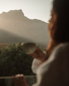 A person in the foreground holds a cup while gazing at a misty mountain landscape through a window at sunrise.