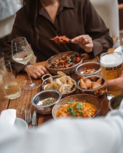 A person in a brown shirt reaches for spicy food from a shared bowl on a wooden table, surrounded by naan, curries, and drinks.