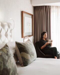 Woman in a dark outfit sits in a lounge chair by a window, holding a teacup and saucer.