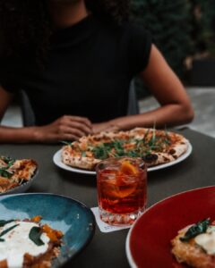 Person in a black shirt sits at an outdoor table with a pizza in front of them and a red cocktail nearby.