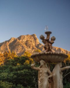 Ornate fountain statue with multiple figures in a garden, mountains in the background at sunset/sunlit.