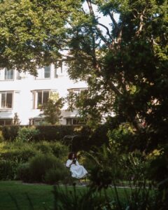 Person in a white dress sits on a bench reading a book in a lush garden with a white apartment building in the background